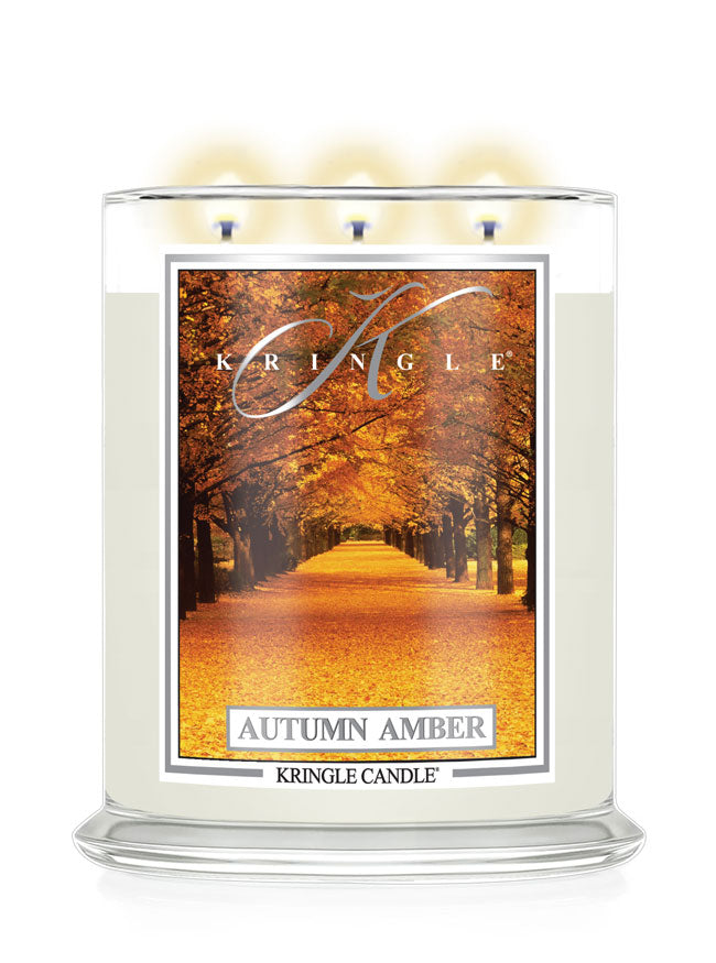 A Kringle Candle in a clear glass jar with three lit wicks, featuring a label that depicts an autumn tree-lined path with orange and yellow leaves. The text 'KRINGLE', 'AUTUMN AMBER', and 'KRINGLE CANDLE' are visible on the label.