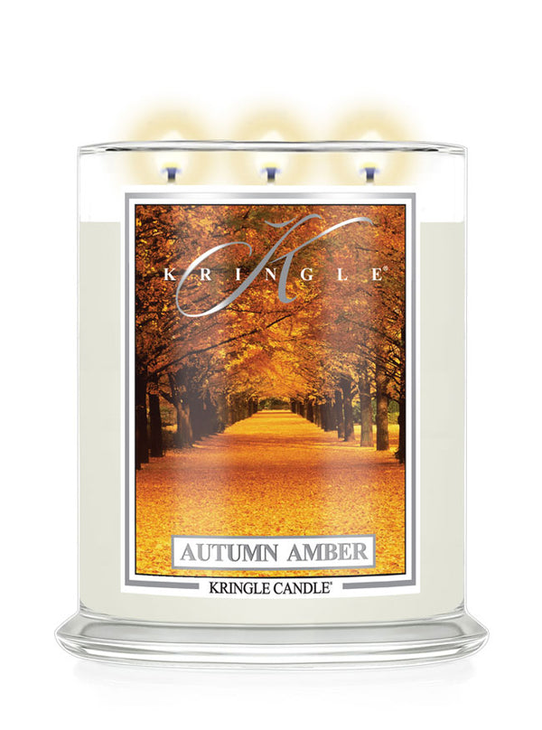 A Kringle Candle in a clear glass jar with three lit wicks, featuring a label that depicts an autumn tree-lined path with orange and yellow leaves. The text 'KRINGLE', 'AUTUMN AMBER', and 'KRINGLE CANDLE' are visible on the label.