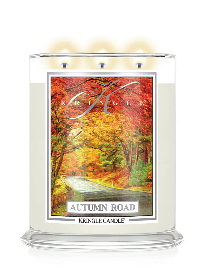 A Kringle Candle in a clear glass jar with three lit wicks, featuring an autumn-themed label that reads 'Autumn Road' and shows a colorful forest scene with red, orange, and yellow foliage.