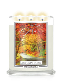 A Kringle Candle in a clear glass jar with three lit wicks, featuring an autumn-themed label that reads 'Autumn Road' and shows a colorful forest scene with red, orange, and yellow foliage.