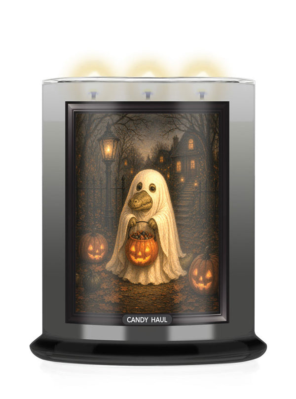 A CANDY HAUL scented candle in a cylindrical glass jar with a black base and a clear top, featuring a Halloween-themed graphic of a ghost holding a jack-o'-lantern on the front label. Three candles are lit at the top, casting a warm glow against a white background.
