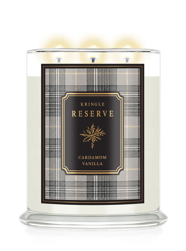 A three-wick scented candle in a clear glass jar with a lit flame, featuring a cream-colored wax and a front label for 'Kringle Reserve Cardamom Vanilla' with a black, gold, and gray plaid pattern.