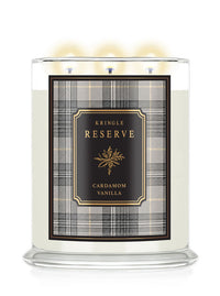 A three-wick scented candle in a clear glass jar with a lit flame, featuring a cream-colored wax and a front label for 'Kringle Reserve Cardamom Vanilla' with a black, gold, and gray plaid pattern.