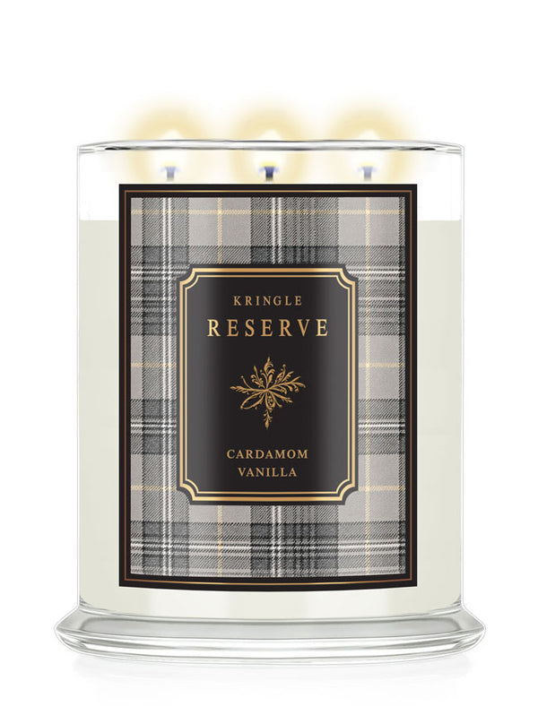 A three-wick scented candle in a clear glass jar with a lit flame, featuring a cream-colored wax and a front label for 'Kringle Reserve Cardamom Vanilla' with a black, gold, and gray plaid pattern.