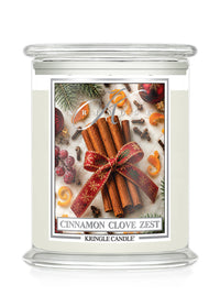 A Kringle Candle jar candle with a clear glass lid and base, filled with off-white wax. The front label features a festive holiday design with cinnamon sticks tied in a red patterned ribbon, cloves, evergreen branches, and citrus slices, with the text 'CINNAMON CLOVE ZEST' and 'KRINGLE CANDLE' visible.