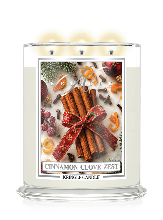 A three-wick Kringle Candle in a clear glass jar with white wax, lit with soft yellow flames. The front label features a festive design with cinnamon sticks tied in a red bow, surrounded by pine needles, cloves, berries, and citrus slices.