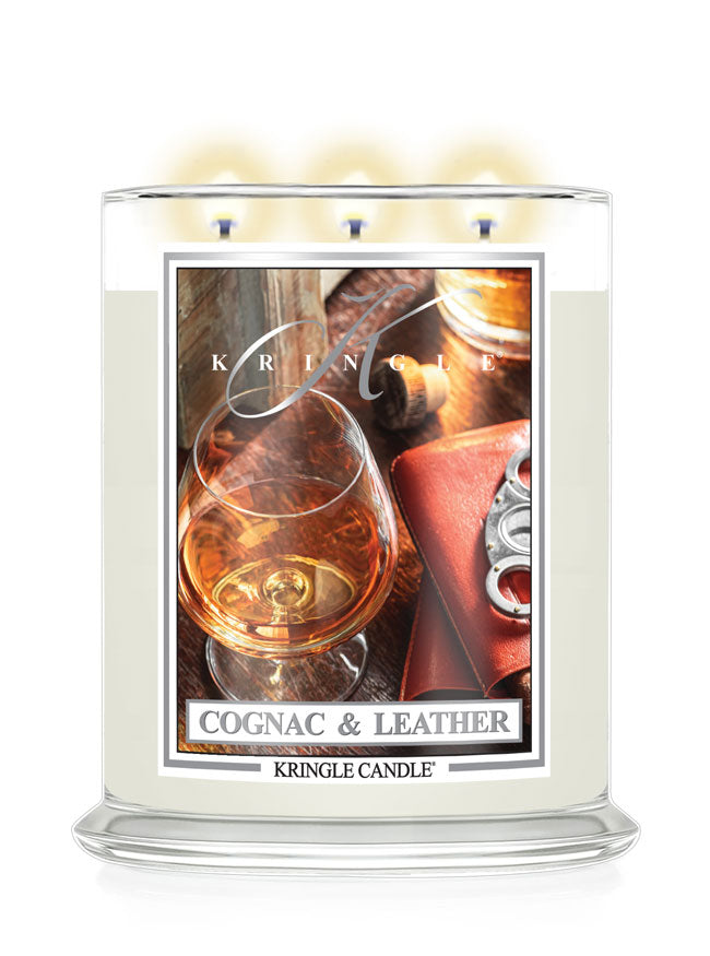 A Kringle Candle in a clear glass jar with three lit wicks, labeled 'Cognac & Leather.' The candle wax is a warm off-white/cream color, and the front label features a photo of a cognac glass, leather, and metallic accents.