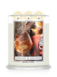 A Kringle Candle in a clear glass jar with three lit wicks, labeled 'Cognac & Leather.' The candle wax is a warm off-white/cream color, and the front label features a photo of a cognac glass, leather, and metallic accents.
