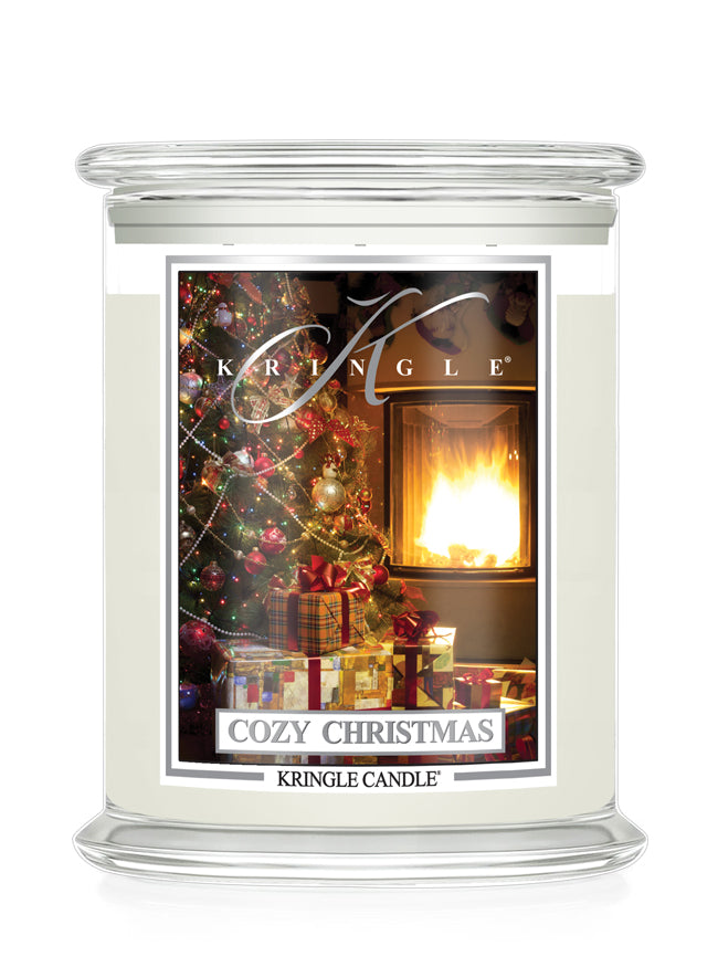 A Kringle Candle jar candle with a clear glass lid and base. The front label features a festive Christmas scene with a decorated tree, wrapped gifts, and a warm fireplace, with the text 'KRINGLE' and 'COZY CHRISTMAS' visible.