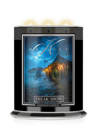 A Kringle Candle Halloween jar labeled 'Freak Show' with a black container, a colorful label showing a circus tent in a blue-toned scene, and three lit wicks at the top on a white background.