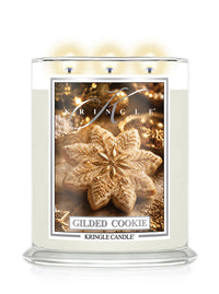 A Kringle Candle in a clear glass jar with three lit wicks, featuring a label that reads 'Gilded Cookie' and shows a decorated cookie on a warm gold holiday background.