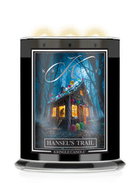 A large black glass jar Halloween candle with three lit wicks, featuring a front label titled 'Hansel's Trail' by Kringle Candle. The label shows a whimsical haunted house surrounded by bare trees at night, with glowing windows and colorful candy decorations.