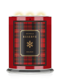 A Kringle Reserve Holiday scented candle in a cylindrical glass jar with a red and black tartan plaid label, featuring a gold snowflake logo and three lit wicks at the top.