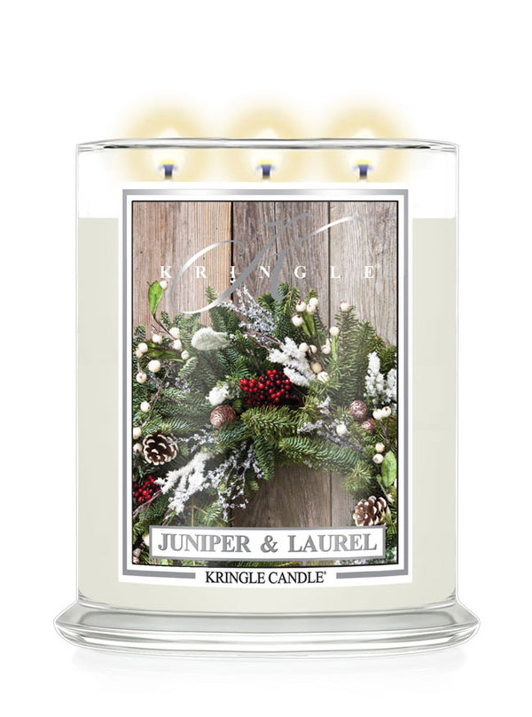 A three-wick Kringle Candle in a clear glass jar with cream-colored wax and a festive winter label reading 'Juniper & Laurel.' The label shows green pine branches, red berries, white frosted details, and a rustic wood background, with the brand name Kringle visible near the top of the label.