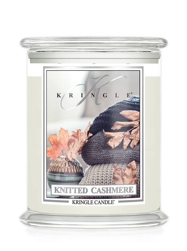 A Kringle Candle in a clear glass jar with a white wax fill and a decorative front label reading 'Knitted Cashmere.' The label shows a dark knit hat, autumn leaves, and a small basket, while the brand name 'Kringle' is visible at the top of the label.