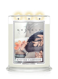 A Kringle Candle in a clear glass jar with three lit wicks, labeled 'Knitted Cashmere.' The candle wax is creamy off-white, and the front label features a cozy knit scene with autumn leaves.