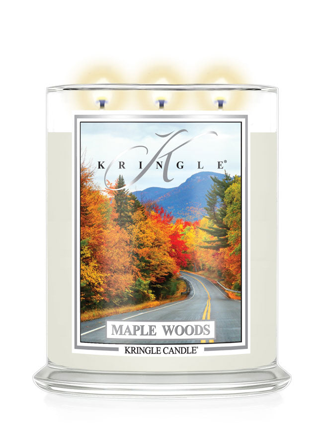 A Kringle Candle in a clear glass jar with three lit wicks, featuring a label that depicts a scenic autumn road through maple woods with mountains in the background., candle labeled Maple Woods.