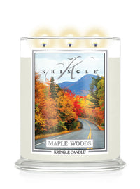 A Kringle Candle in a clear glass jar with three lit wicks, featuring a label that depicts a scenic autumn road through maple woods with mountains in the background., candle labeled Maple Woods.