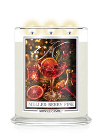 A Kringle Candle in a clear glass jar with three lit wicks, featuring a front label for 'Mulled Berry Pine' with an illustration of a berry drink, orange slices, pinecones, and warm holiday lights.