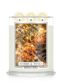 A three-wick Kringle Candle in a clear glass jar with cream-colored wax, featuring a festive winter label that reads 'MYRRH & TONKA' and shows warm spices, evergreen branches, and glowing lights.