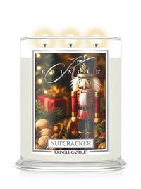 A large three-wick holiday candle in a clear glass jar with a white wax body and a festive front label featuring a nutcracker, ornaments, and the text 'Kringle' and 'Nutcracker' by Kringle Candle.
