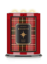 A Kringle Reserve 'Silent Night' scented candle in a red glass jar with a black and red tartan label, featuring three lit wicks and a clear glass base.