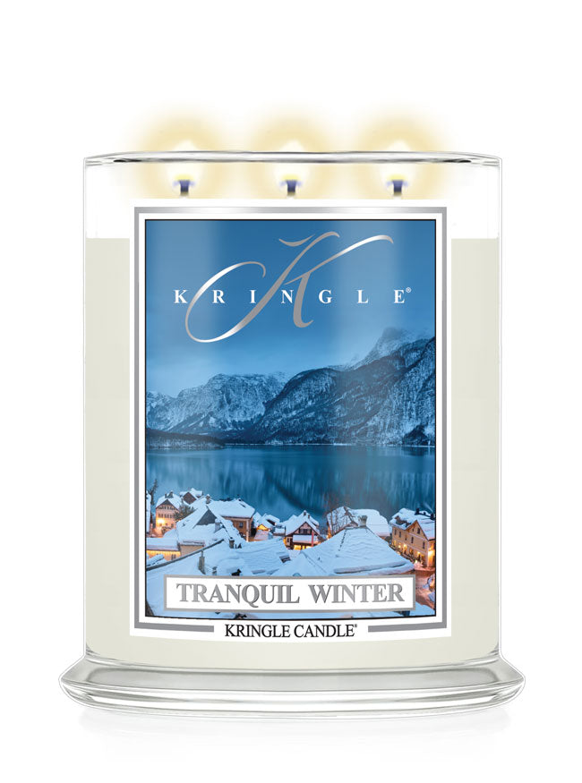 A Kringle Candle in a clear glass jar with three lit wicks, featuring a winter landscape label that reads 'Tranquil Winter' and shows snowy rooftops, mountains, and a blue lake.
