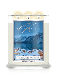 A Kringle Candle in a clear glass jar with three lit wicks, featuring a winter landscape label that reads 'Tranquil Winter' and shows snowy rooftops, mountains, and a blue lake.