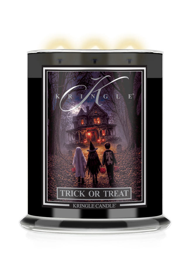 A Kringle Candle 'Trick or Treat' scented candle in a tall black glass jar with three lit wicks at the top, featuring a Halloween-themed label illustration of children facing a haunted house.