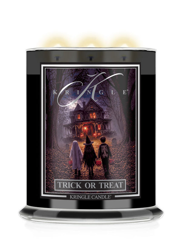 A Kringle Candle 'Trick or Treat' scented candle in a tall black glass jar with three lit wicks at the top, featuring a Halloween-themed label illustration of children facing a haunted house.
