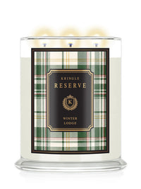 A three-wick scented candle in a clear glass jar with lit flames, featuring a green and white plaid label with a black center panel that reads 'KRINGLE RESERVE WINTER LODGE' in gold lettering.