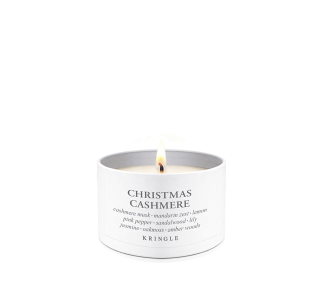 A lit white scented candle in a white cylindrical tin with a single flame visible at the top. The label reads 'CHRISTMAS CASHMERE' with smaller scent notes and the brand 'KRINGLE' at the bottom, set against a plain white background.