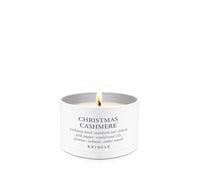 A lit white scented candle in a white cylindrical tin with a single flame visible at the top. The label reads 'CHRISTMAS CASHMERE' with smaller scent notes and the brand 'KRINGLE' at the bottom, set against a plain white background.