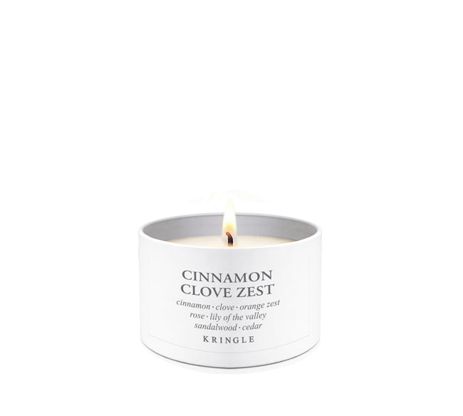 A lit white candle in a cylindrical white tin with the text 'CINNAMON CLOVE ZEST' and 'KRINGLE' printed on the front, set against a solid white background.