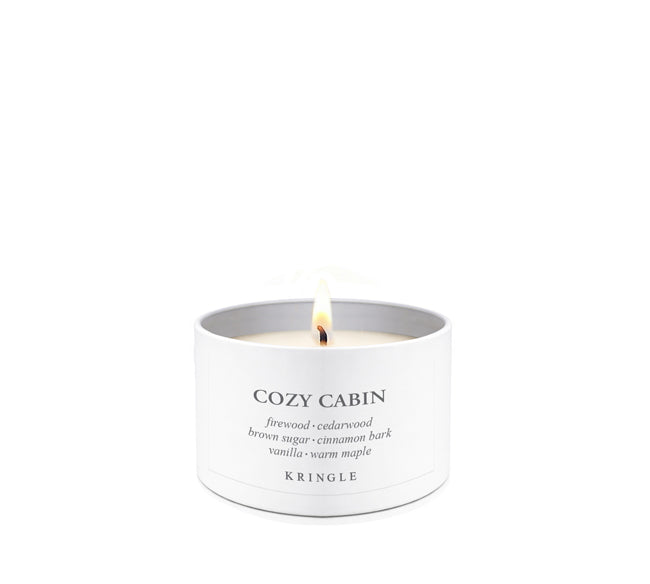 A lit white candle in a white cylindrical tin with the brand name KRINGLE and scent name COZY CABIN printed on the front, set against a seamless white background.
