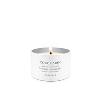A lit white candle in a white cylindrical tin with the brand name KRINGLE and scent name COZY CABIN printed on the front, set against a seamless white background.