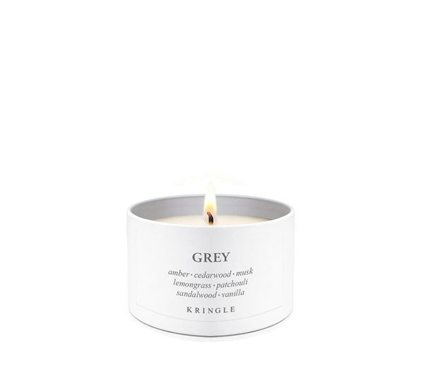 A lit white candle in a cylindrical white metal tin with the brand name KRINGLE visible on the front. The label reads 'GREY' with scent notes including amber, cedarwood, mistel, lemon grass, patchouli, sandalwood, and vanilla.