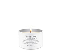 A lit white candle in a white cylindrical tin with the text 'KNITTED CASHMERE' and 'KRINGLE' printed on the front, set against a solid white background.