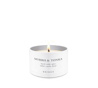 A lit white candle in a white cylindrical tin container with the text 'MYRRH & TONKA' and 'KRINGLE' printed on the front, set against a seamless white background.