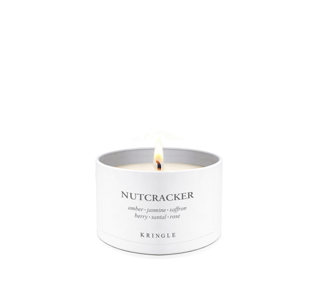 A lit white candle in a white cylindrical tin container with the scent name 'NUTCRACKER' and brand 'KRINGLE' printed on the front, set against a solid white background.