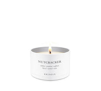 A lit white candle in a white cylindrical tin container with the scent name 'NUTCRACKER' and brand 'KRINGLE' printed on the front, set against a solid white background.
