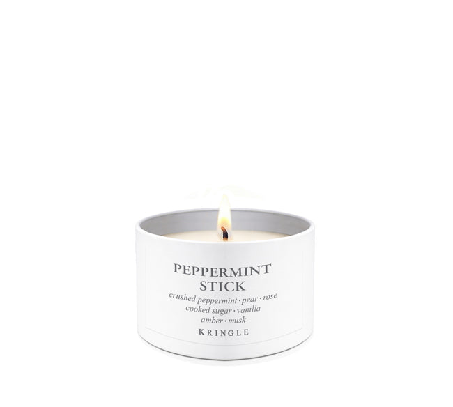 A lit Kringle Peppermint Stick candle in a white cylindrical tin with a single flame visible, centered on a clean white background.