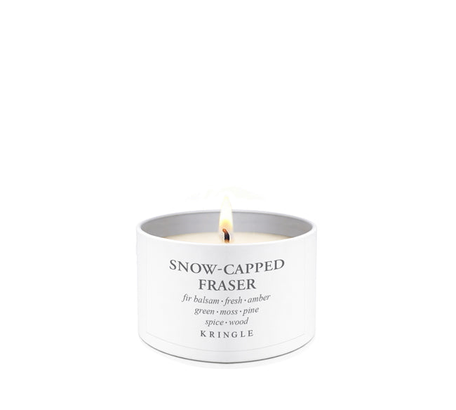 A lit white candle in a cylindrical white tin with the text 'SNOW-CAPPED FRASER' and 'KRINGLE' printed on the front, set against a solid white background.