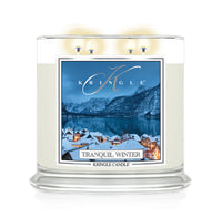 A Kringle Candle in a clear glass jar with three lit wicks, featuring a winter landscape label that reads 'Tranquil Winter' and shows snowy rooftops, mountains, and a blue lake.