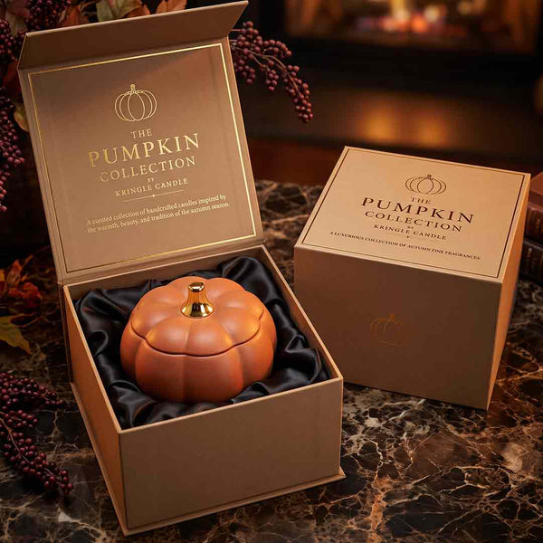 A pumpkin-shaped candle in an open brown gift box with gold lettering, shown beside another closed box on a marble surface with decorative berries in the background. The candle is orange-brown with a small gold top knob.