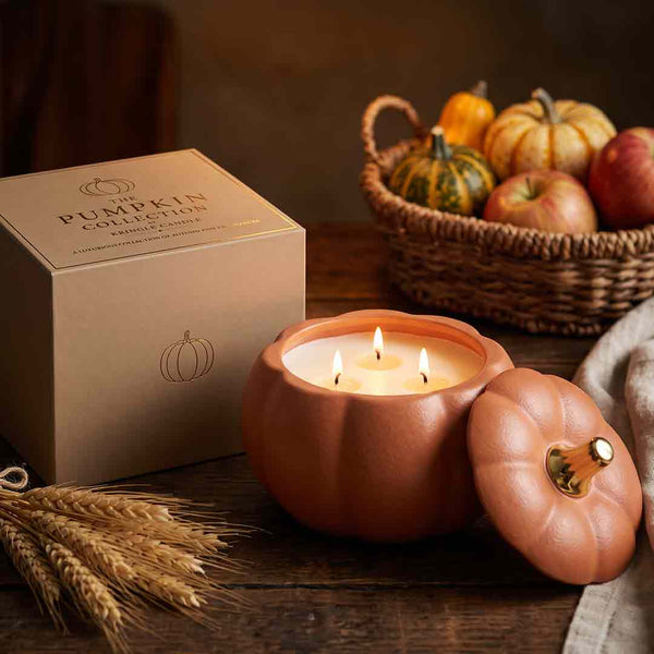 A lit pumpkin-shaped candle with its lid beside it on a rustic wooden table, next to a kraft box labeled 'Pumpkin Collection' and autumn props including wheat stalks and a basket of pumpkins and apples.
