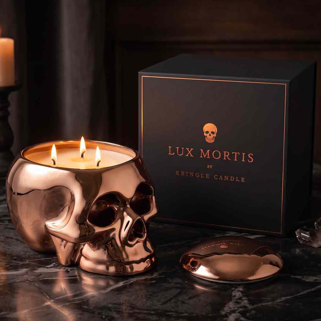 A skull-shaped copper candle with three lit wicks sits on a dark marble surface beside its black branded box and the detached lid. Visible text on the box reads 'LUX MORTIS BY KRINGLE CANDLE'.