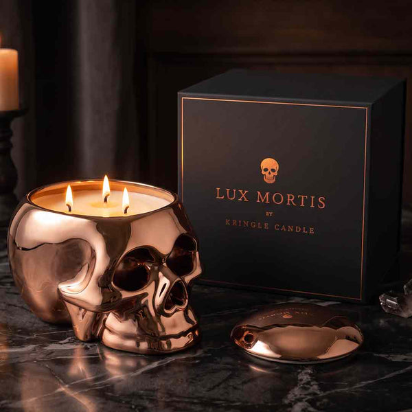 A skull-shaped copper candle with three lit wicks sits on a dark marble surface beside its black branded box and the detached lid. Visible text on the box reads 'LUX MORTIS BY KRINGLE CANDLE'.