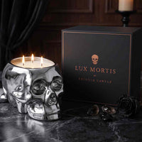 A skull-shaped silver candle holder with three lit wicks sits beside a black branded box reading 'LUX MORTIS BY KRINGLE CANDLE' on a dark marble surface.