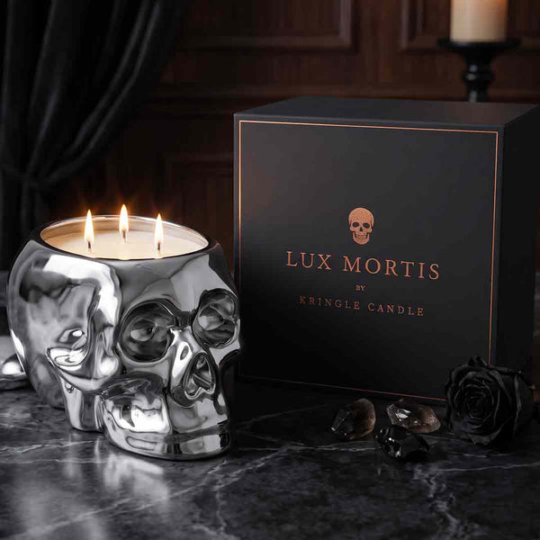 A skull-shaped silver candle holder with three lit wicks sits beside a black branded box reading 'LUX MORTIS BY KRINGLE CANDLE' on a dark marble surface.